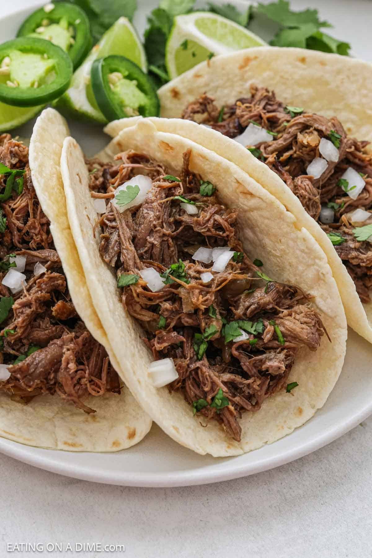 Three beef barbacoa tacos in flour tortillas, topped with chopped onions and cilantro, served with sliced jalapeños, lime wedges, and cilantro—just like a slow cooker beef carnitas recipe brought to life on a white plate.