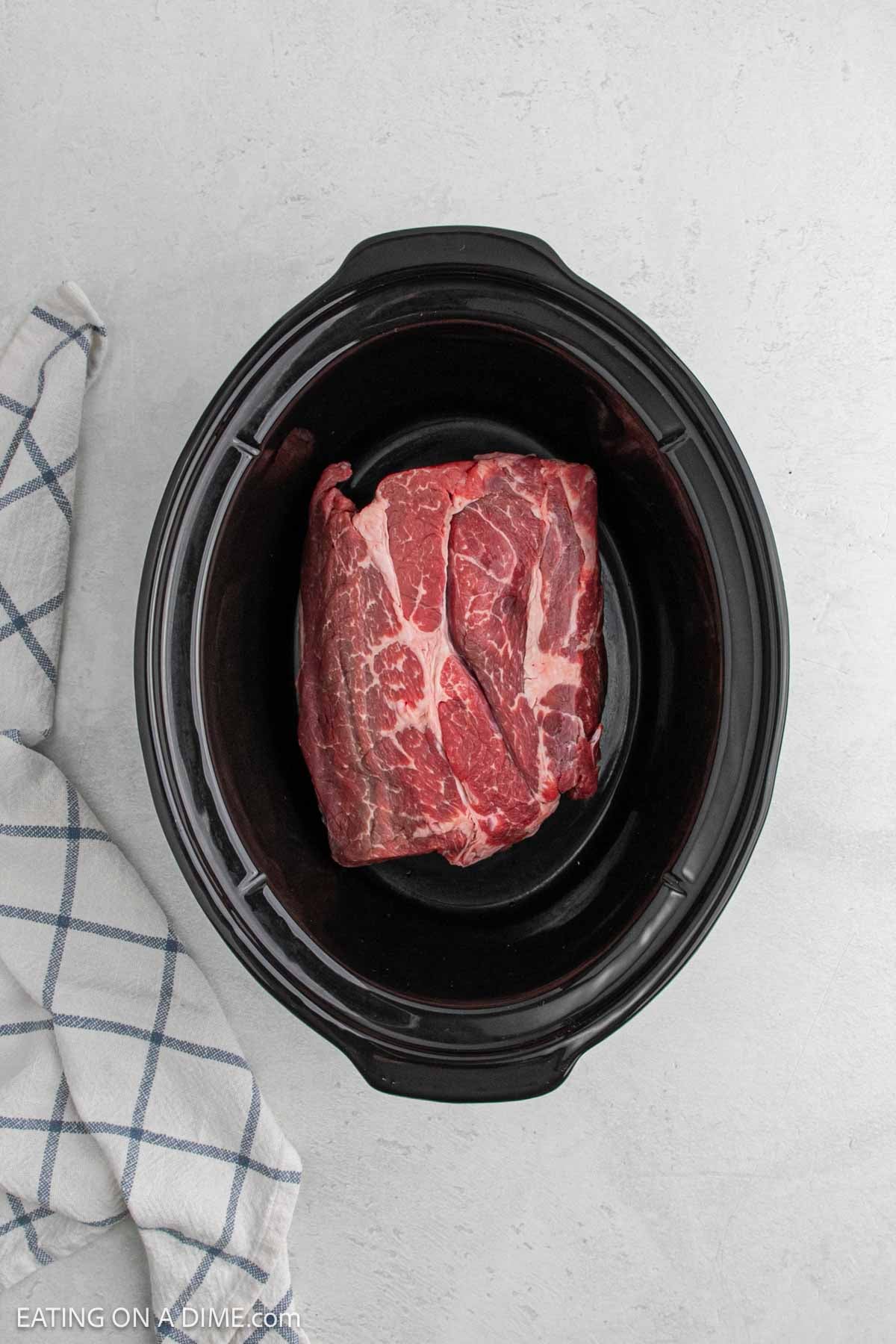 A raw beef roast sits in a black slow cooker on a light countertop, ready to become a delicious slow cooker beef carnitas recipe, with a white and blue checkered kitchen towel nearby.