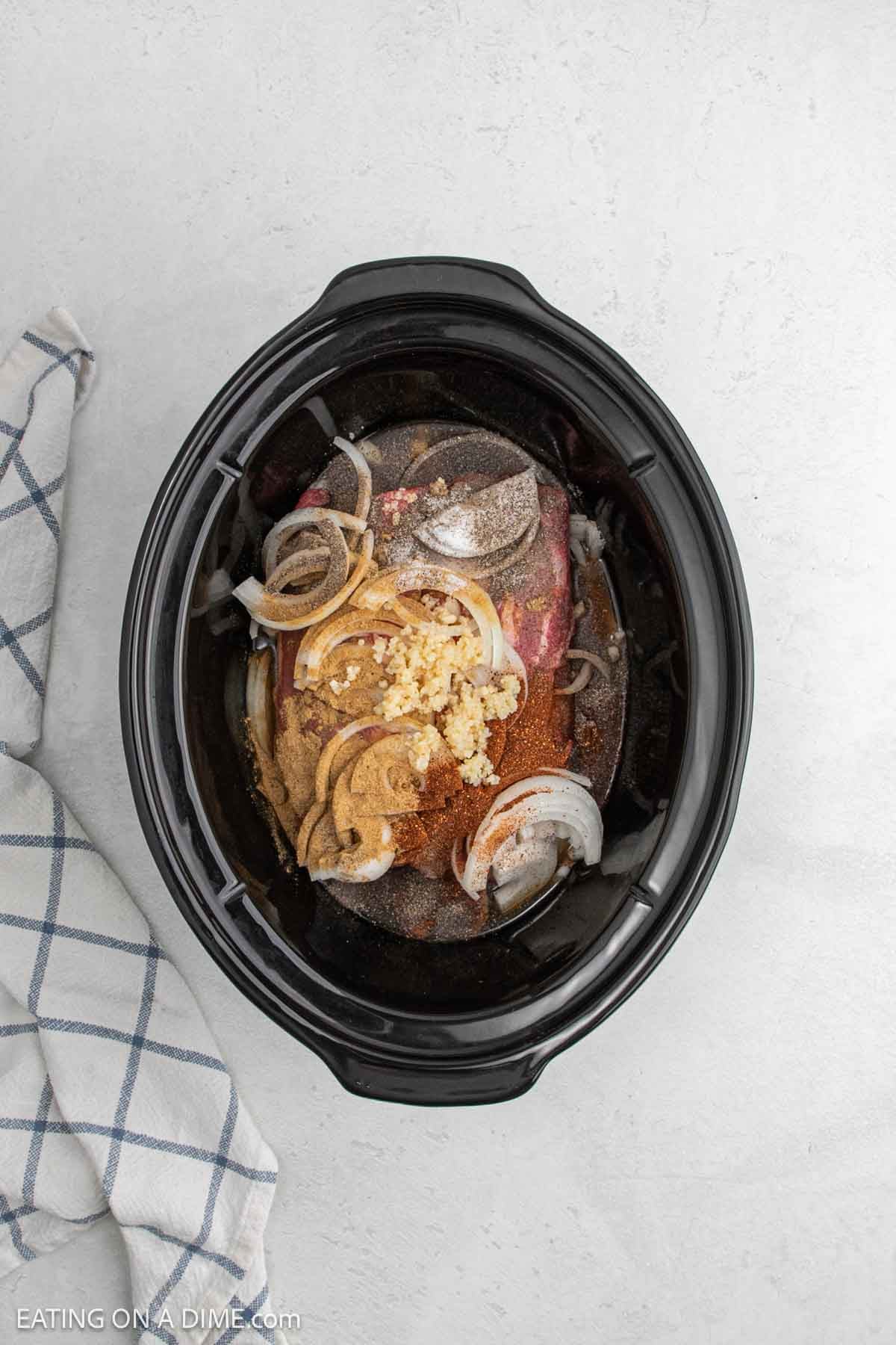 A black slow cooker filled with sliced onions, minced garlic, mustard, seasonings, and raw meat sits on a light countertop next to a white and blue checkered towel—perfect for an easy slow cooker beef carnitas recipe.