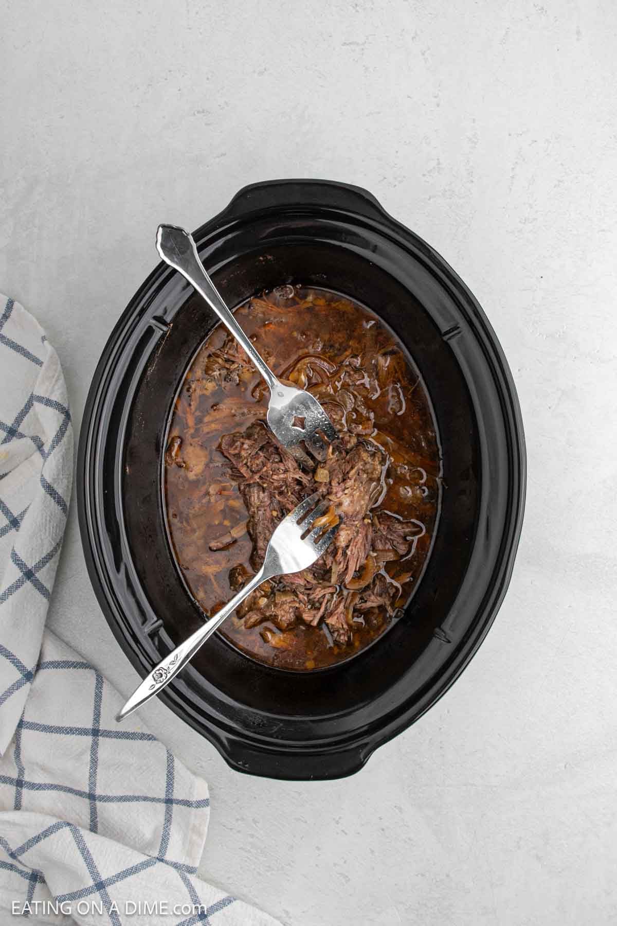Shredded beef being pulled apart with two forks in a slow cooker filled with rich brown sauce, next to a blue and white checkered towel—perfect for your favorite slow cooker beef carnitas recipe.