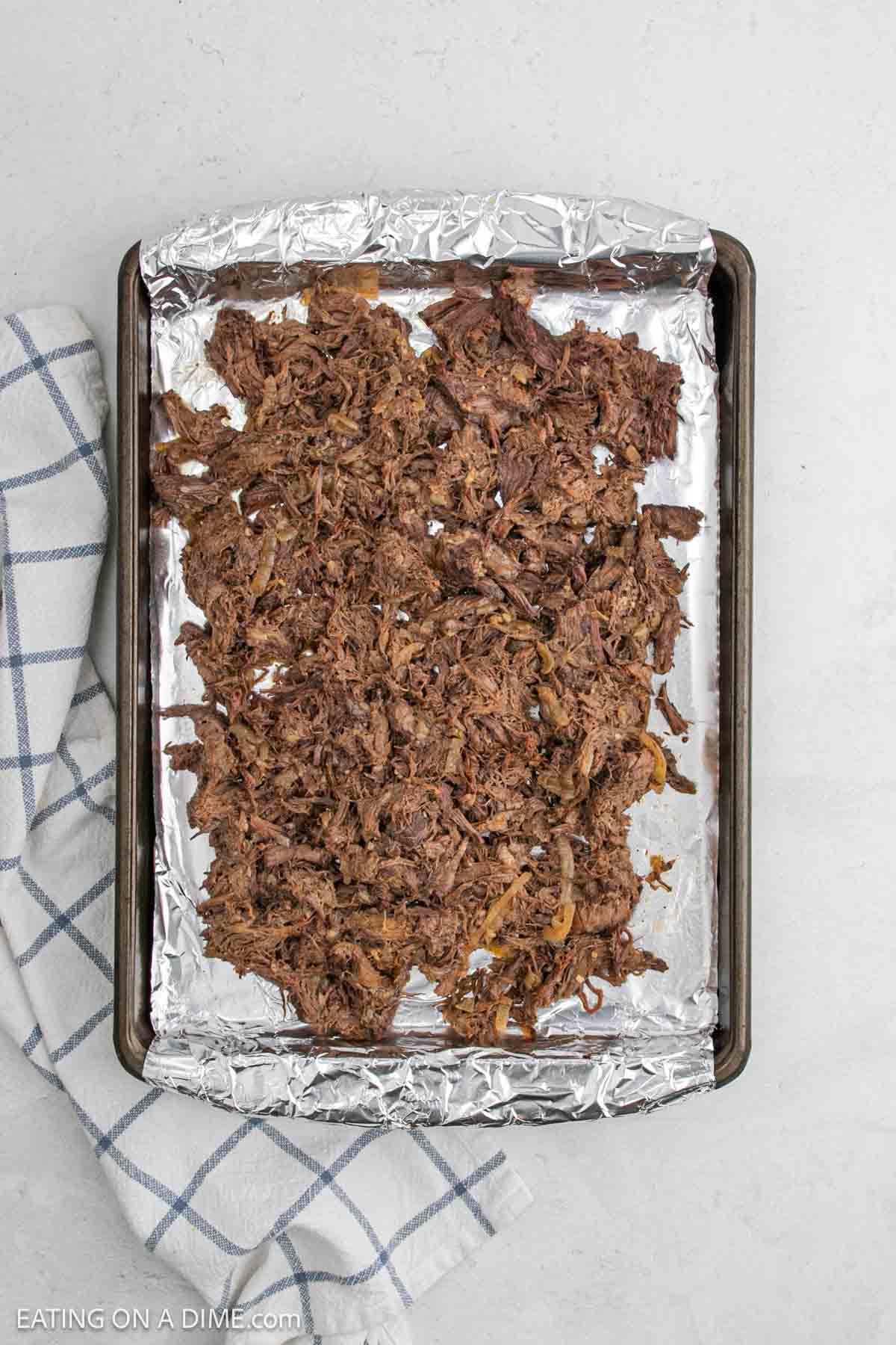 Shredded slow cooker beef carnitas recipe spread on a foil-lined baking sheet, ready to be broiled, with a white and blue checkered kitchen towel beside the tray on a light surface.