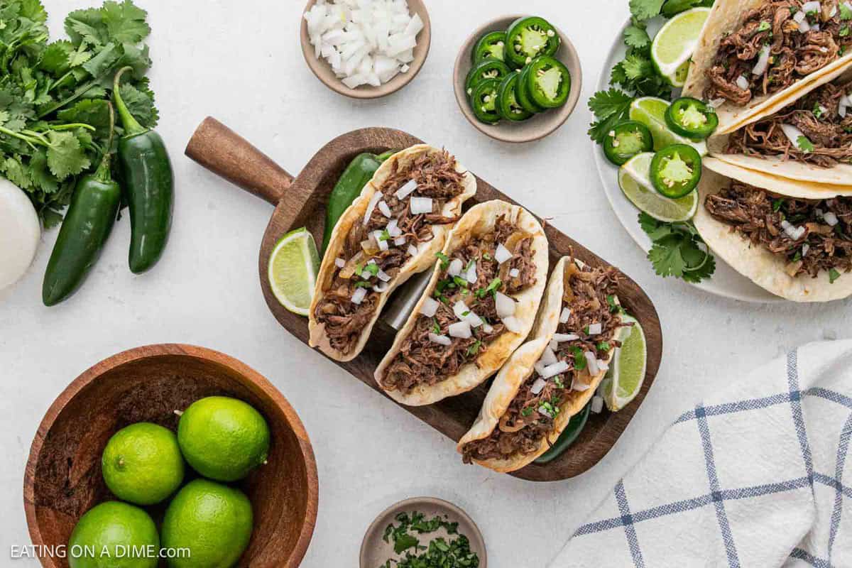 Three tacos filled with slow cooker beef carnitas, topped with chopped onions and cilantro, served on a cutting board with lime wedges. Fresh limes, jalapeños, cilantro, and bowls of toppings surround the board.
