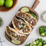 Three shredded beef tacos, made using a slow cooker beef carnitas recipe, are topped with chopped onions and cilantro on a wooden tray with lime wedges. Sliced jalapeños, chopped onions, and whole limes sit in small bowls nearby.