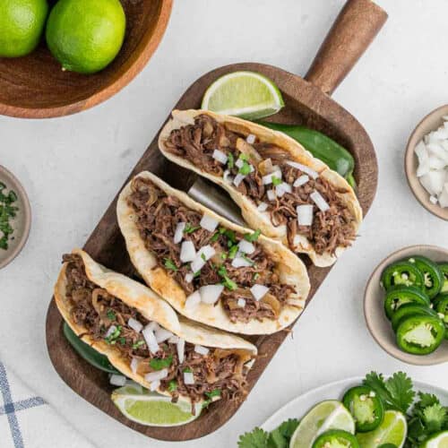 Three shredded beef tacos, made using a slow cooker beef carnitas recipe, are topped with chopped onions and cilantro on a wooden tray with lime wedges. Sliced jalapeños, chopped onions, and whole limes sit in small bowls nearby.