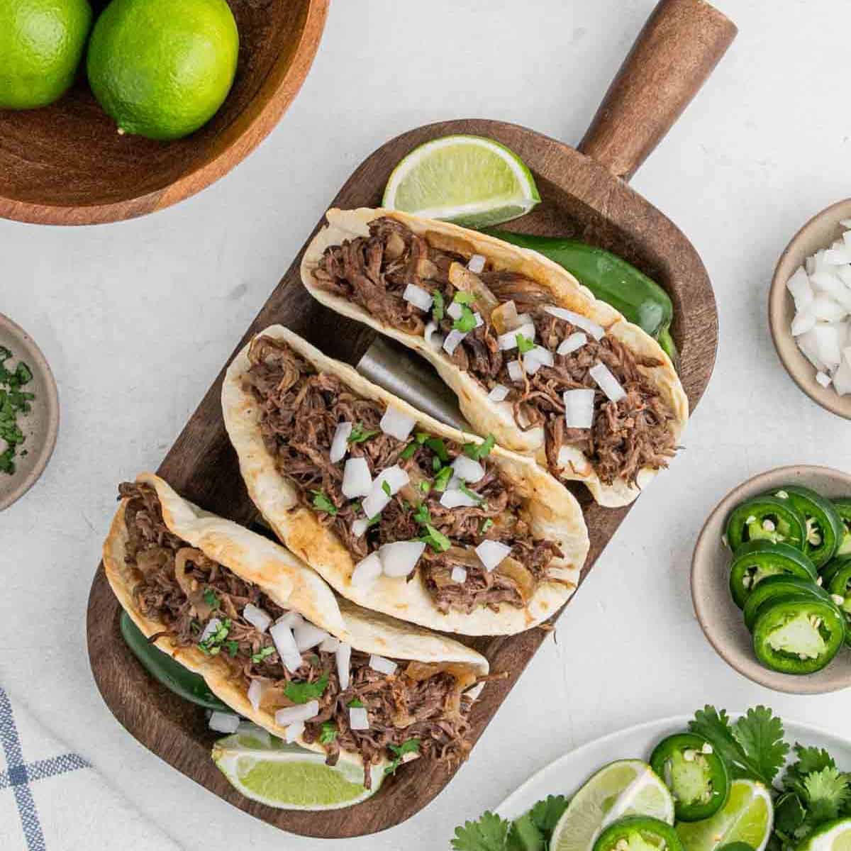 Three shredded beef tacos, made using a slow cooker beef carnitas recipe, are topped with chopped onions and cilantro on a wooden tray with lime wedges. Sliced jalapeños, chopped onions, and whole limes sit in small bowls nearby.