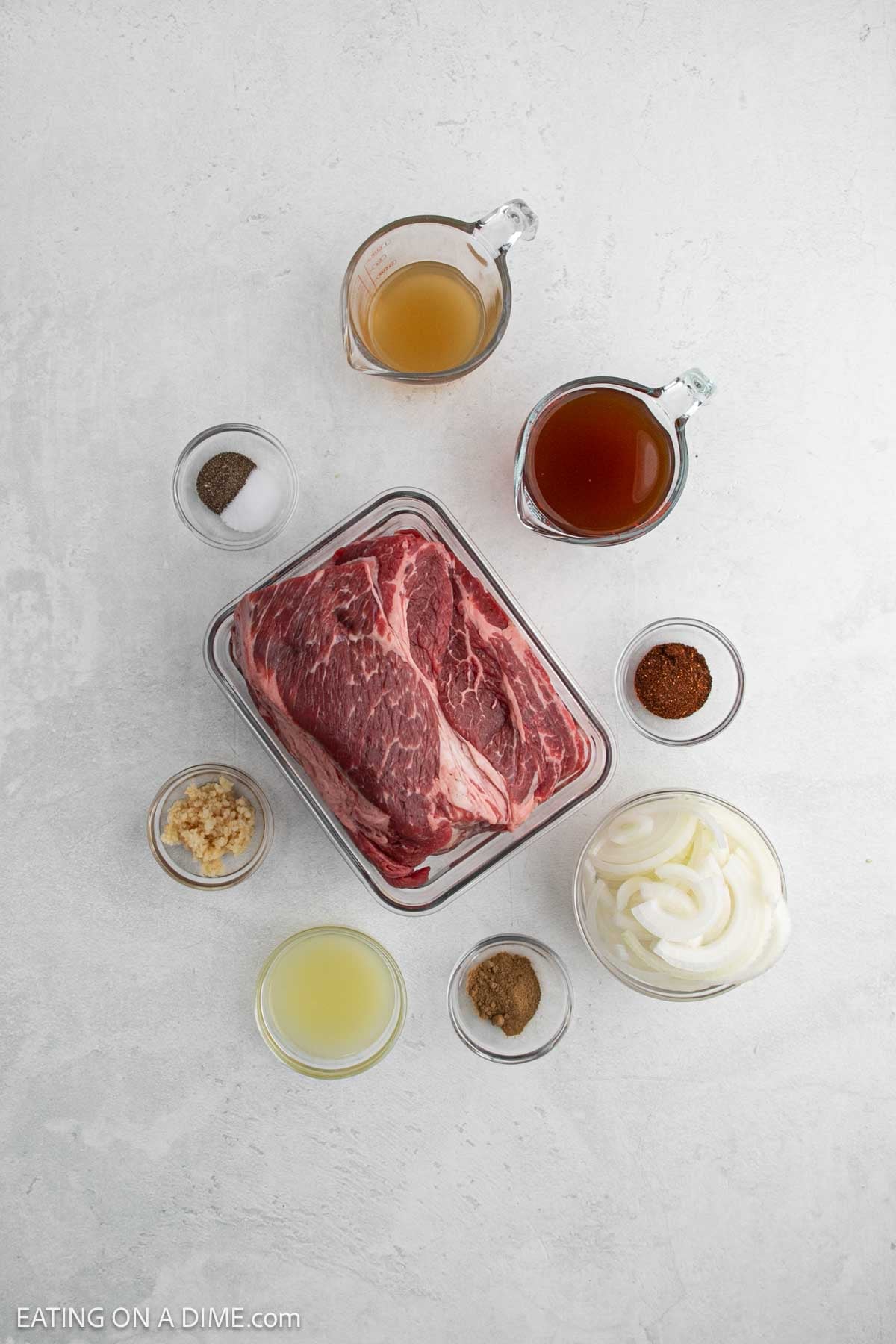 A raw chuck roast in a square container is surrounded by small bowls of sliced onions, minced garlic, ground spices, black pepper, juice, and two glass measuring cups with broth—perfect ingredients for a slow cooker beef carnitas recipe.