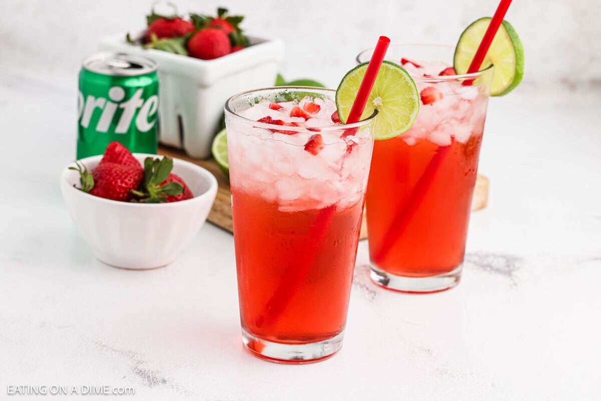 Two glasses of Strawberry Limeade that have a lime wedge on the rim with bowls of fresh strawberries in the background. 