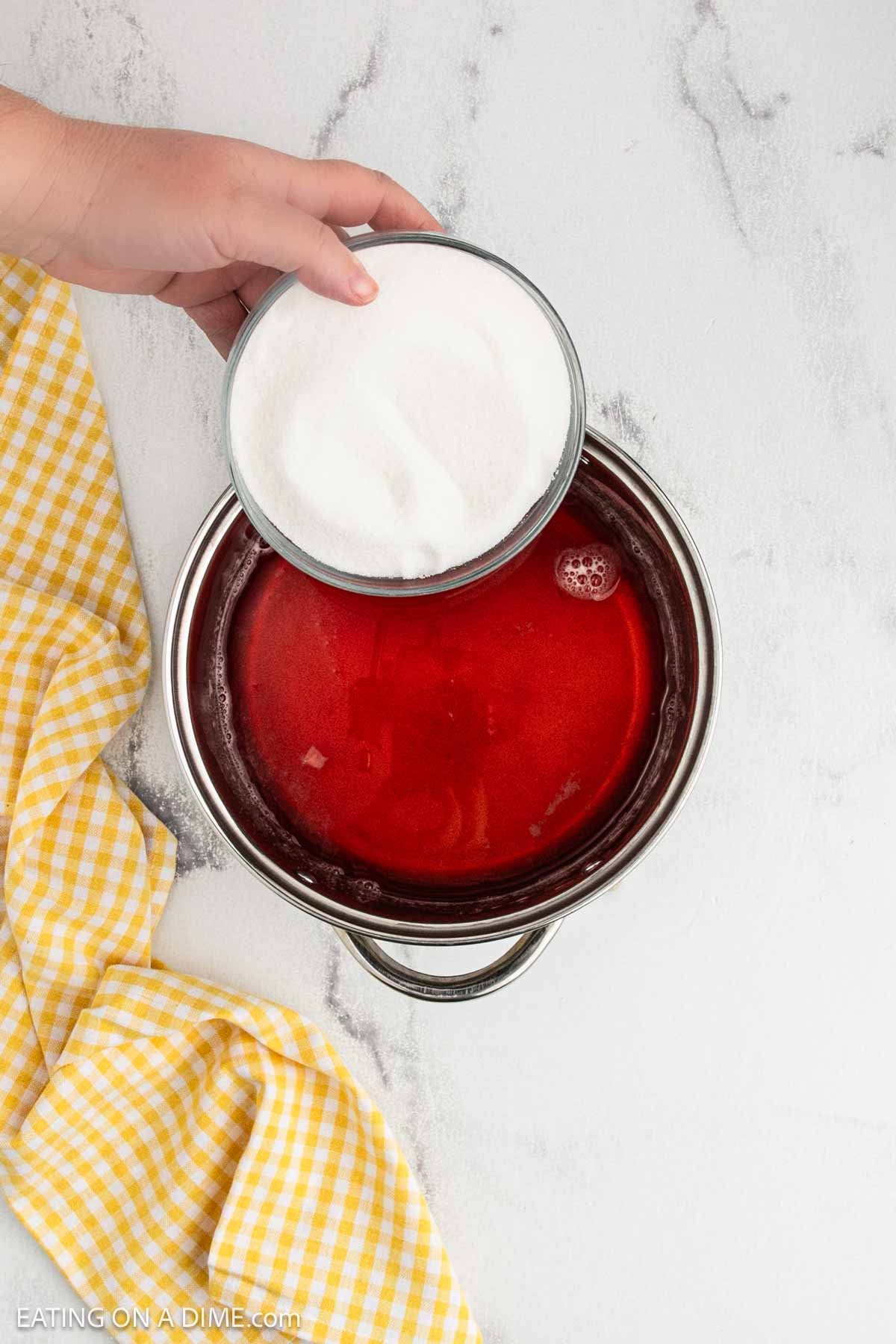 Pouring a bowl of white sugar into the large pot of red liquid.