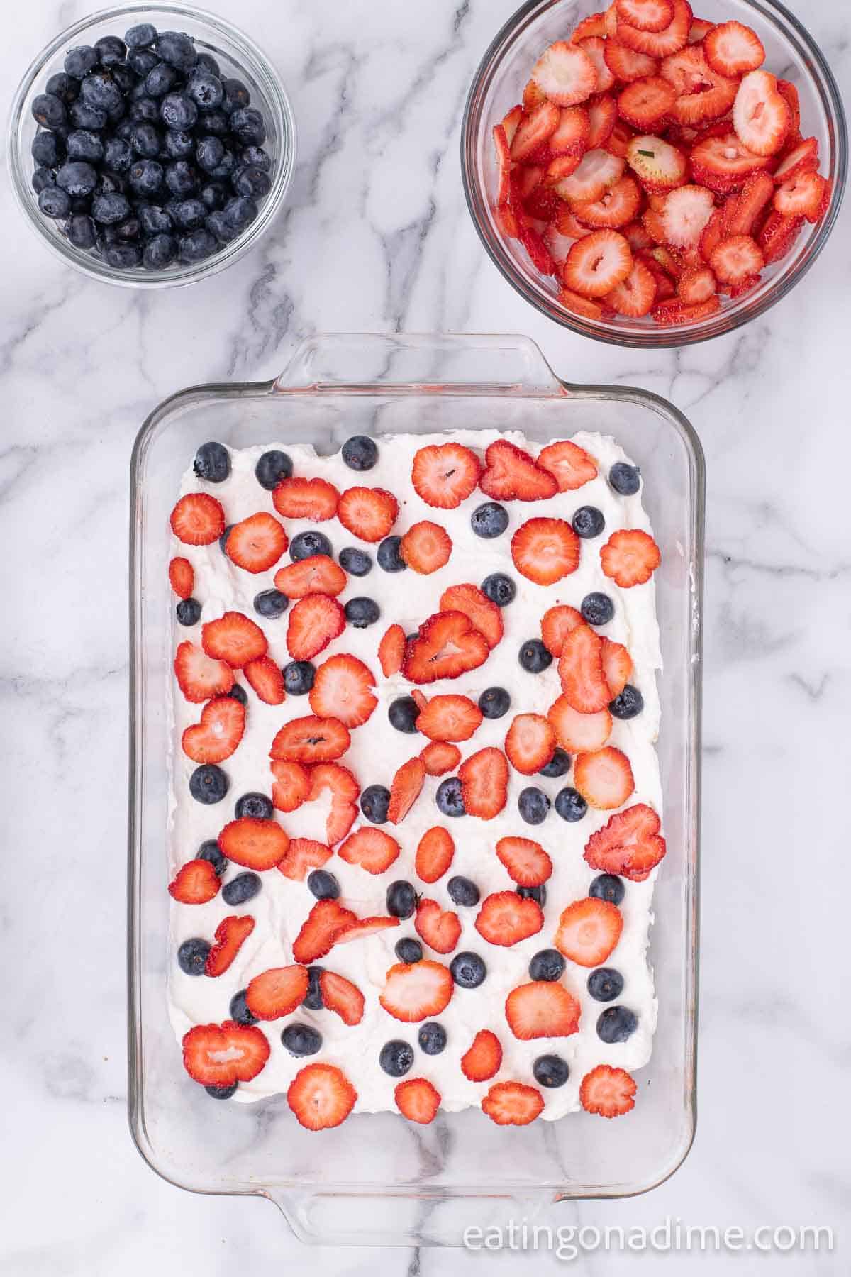 Strawberries and blueberries topped on the cream layer in the baking dish with small bowls of blueberries and strawberries on the side