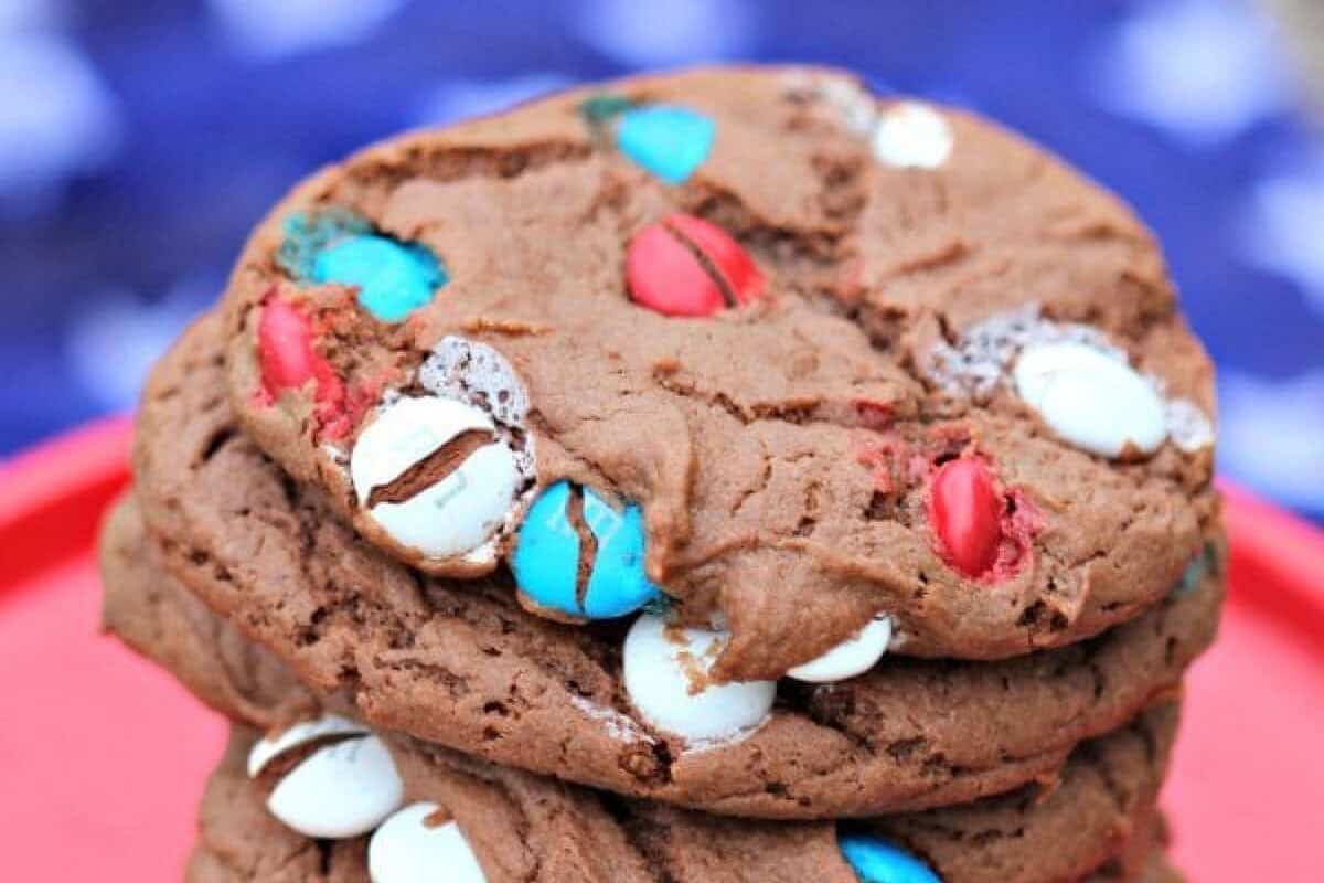 You only need 5 ingredients to make these delicious 4th of July Cake Mix Cookies. These cake mix cookies are one of the best 4th of July desserts. A stack of 4th of July Cookies with red, white, and blue candy-coated chocolates sits on a red surface, against a blurry blue and white star-patterned background.