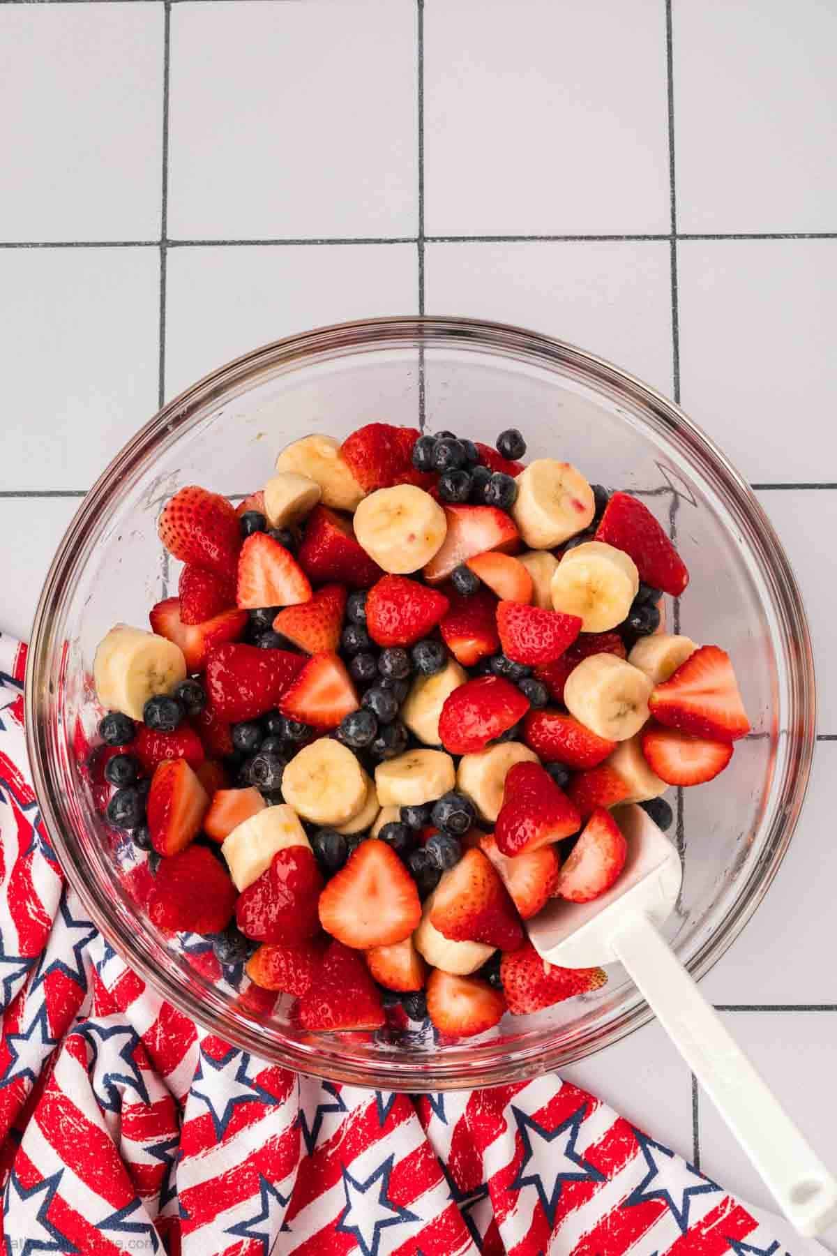 Slice strawberries, bananas, and blueberries combined in a bowl with a spatula with the dressing