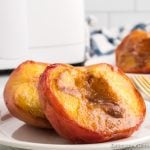 Two baked peach halves with caramelized centers sit on a white plate. A fork rests beside these delicious Air Fryer Peaches, with a blurred kitchen background visible.