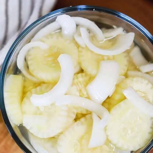 A glass bowl filled with sliced cucumbers and onions in a light brine. The cucumber and onion slices are layered, showcasing their translucent appearance. This image highlights a cucumber onion salad recipe preparation.
