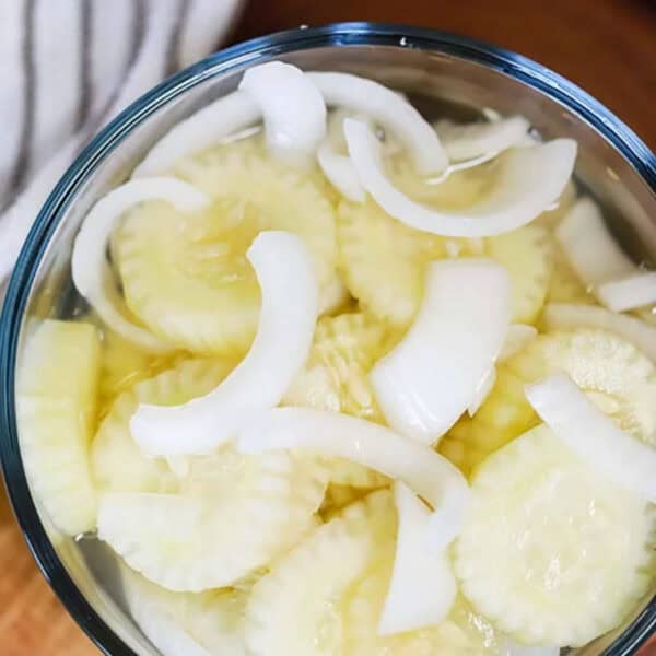 A glass bowl filled with sliced cucumbers and onions in a light brine. The cucumber and onion slices are layered, showcasing their translucent appearance. This image highlights a cucumber onion salad recipe preparation.