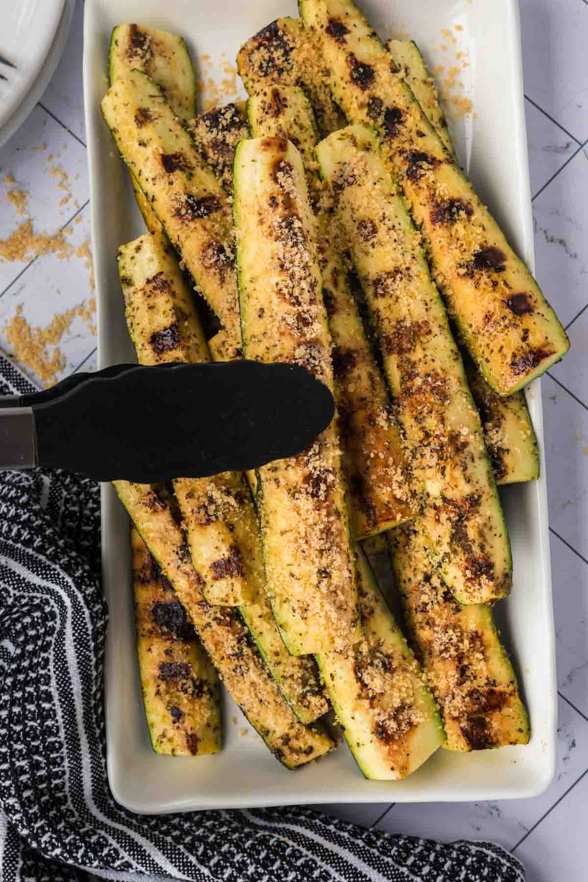 Parmesan Zucchini Spears stacked on a platter with a pair of tongs holding a zucchini spears ready to serve