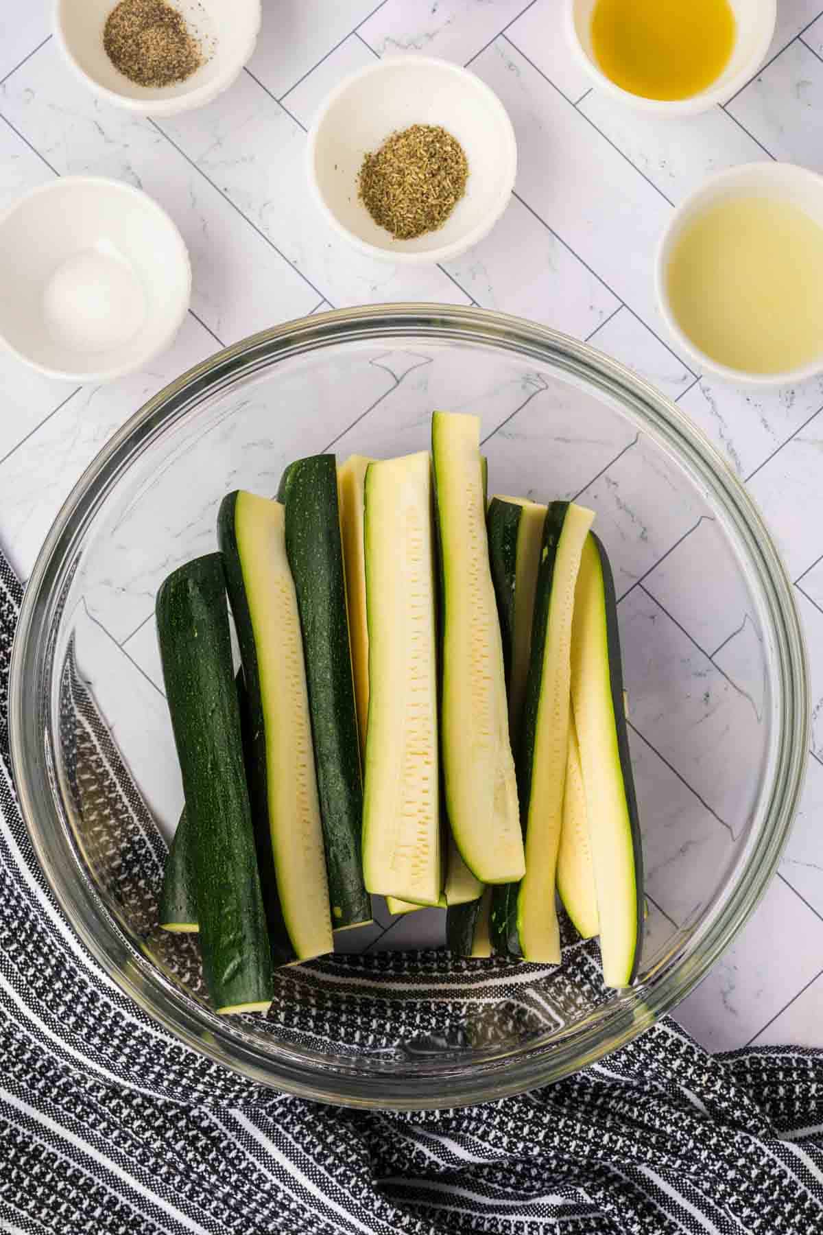 Zucchini spears in a clear bowl with small bowls of seasoning and oil