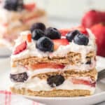 A close-up of a 4th of July Icebox Cake with graham crackers, whipped cream, sliced strawberries, and whole blueberries, served on a white plate.
