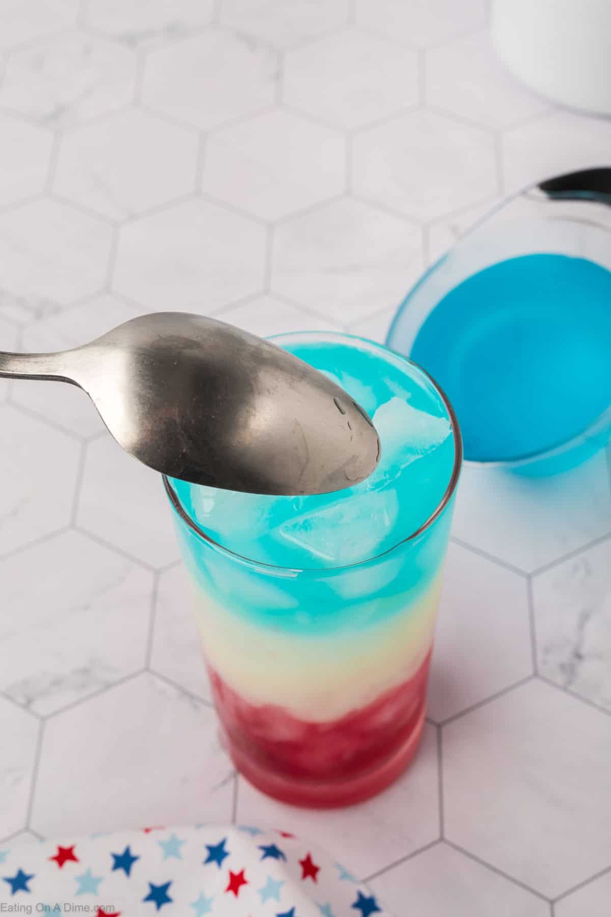 Pouring the blue gatorade on top of the pina colada layer and the red juice in a clear glasses with ice