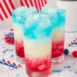 Three tall glasses filled with a festive Red White and Blue Drink, each layered and topped with ice, sit among patriotic decorations and American flags in the background.