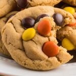 A close-up of a thick, soft Reese's Pieces Cookies studded with colorful candy-coated chocolates, with a bite taken out of it, resting on a white plate.