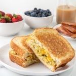 A Scrambled Egg Sandwich with melted cheese is cut in half on a white plate, accompanied by bowls of strawberries and blueberries, crispy bacon on a plate, and a glass of iced coffee in the background.