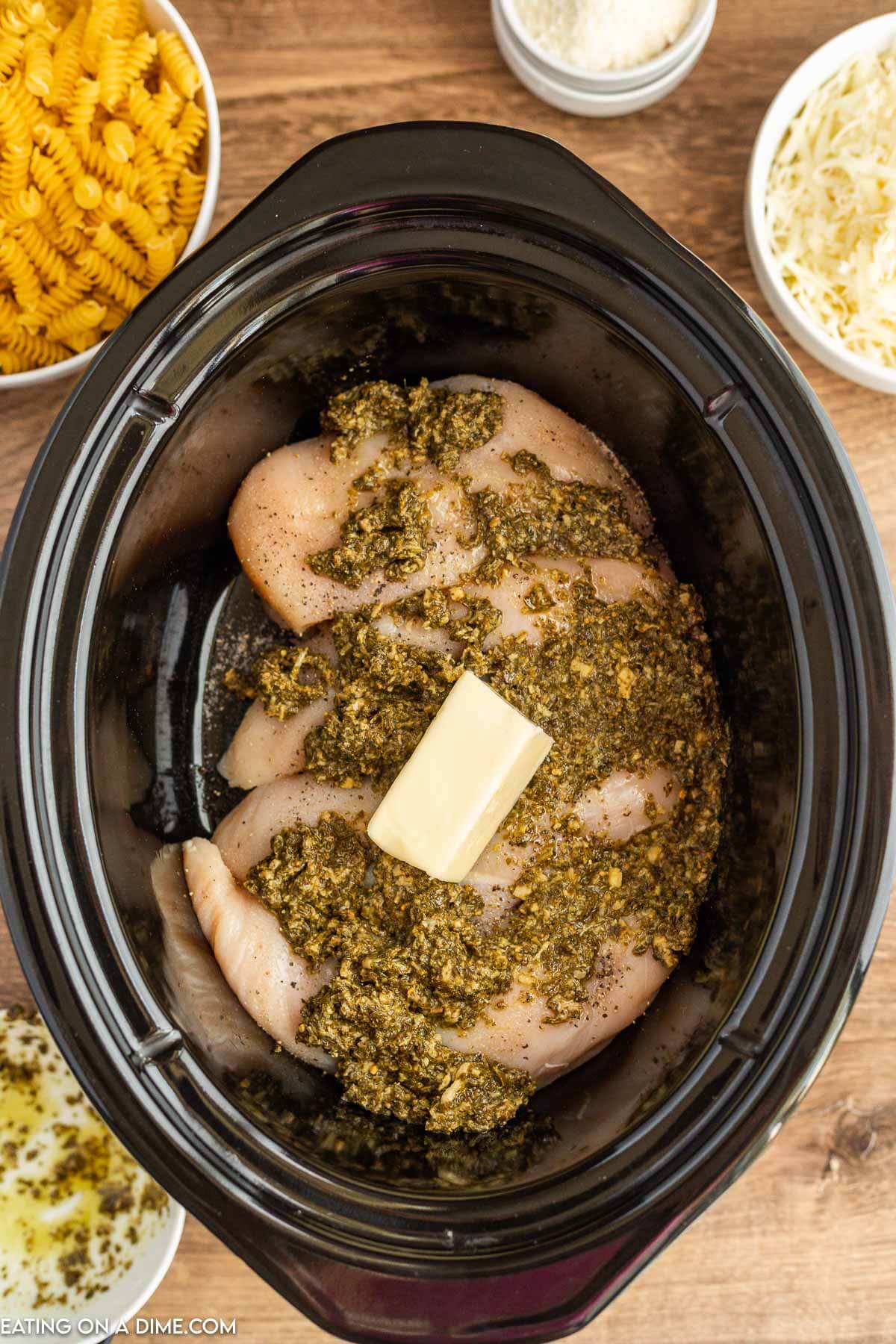 Placing the chicken breast in the slow cooker and topping with butter and pesto
