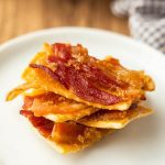 A stack of crispy bacon brittle pieces, reminiscent of savory bacon crackers with a golden, caramelized coating, sits on a white plate, with a blurred checkered cloth in the background.