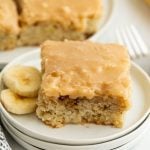 A square piece of banana cake with light brown frosting on a white plate, next to banana slices. Reminiscent of banana bread brownies, another piece of cake and two forks are visible in the background.