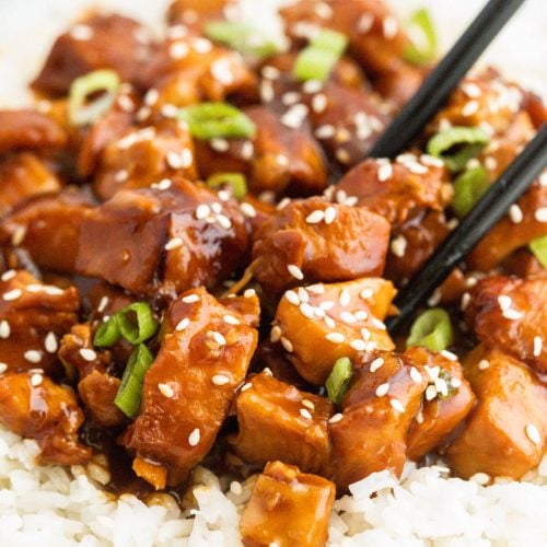 Cubes of glazed teriyaki chicken with sesame seeds and chopped green onions served over white rice, with a pair of black chopsticks picking up a piece—perfect for fans of instant pot orange chicken flavors.