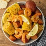 An instant pot shrimp boil fills the bowl with shrimp, corn on the cob, sausage slices, red potatoes, and lemon wedges, all garnished with herbs. It sits on a wooden table beside a checkered cloth and slices of bread.