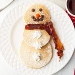 Three round snowman pancakes are arranged on a white plate, with chocolate chips and an orange candy for the face, whipped cream buttons, a bacon scarf, and a dusting of powdered sugar on top.