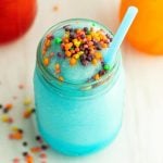 A mason jar filled with a blue Sonic Nerds Slushie, topped with colorful candy bits and a blue straw. Blurred red and orange drinks are visible in the background, with some candy pieces scattered on the white surface.