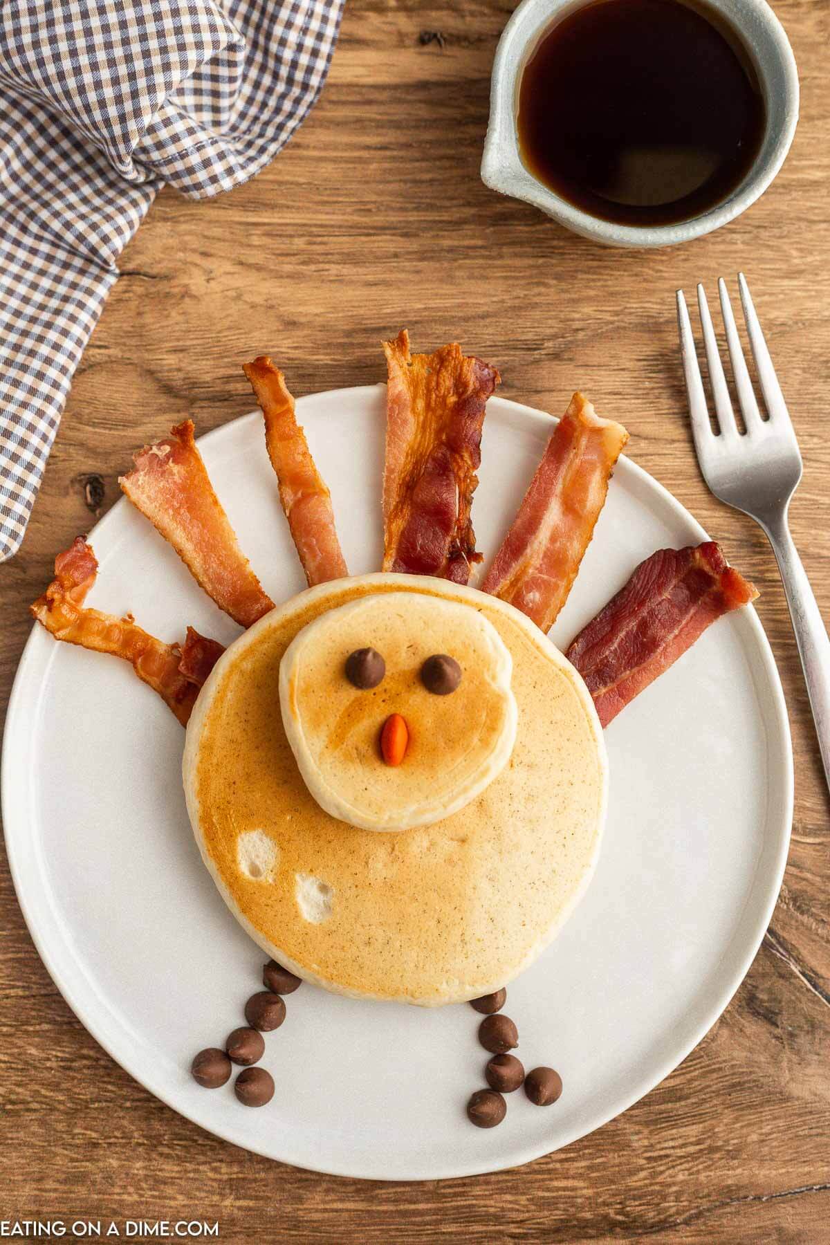 Turkey shaped pancake on a plate with strips of bacon as the turkey feathers and topped with chocolate and orange M&M as the nose & eyes