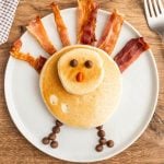Turkey Shaped Pancakes on a plate with bacon strips as the feathers with chocolate chips as the eyes and legs with orange M&M as the nose.