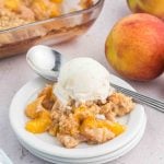 A serving of peach crumble cobbler topped with a scoop of vanilla ice cream sits on a small white plate with a spoon, with fresh peaches and a baking dish in the background.