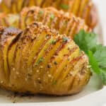 Close-up of golden-brown Hasselback Potatoes, thinly sliced and seasoned with herbs and spices, garnished with fresh parsley on a white plate.