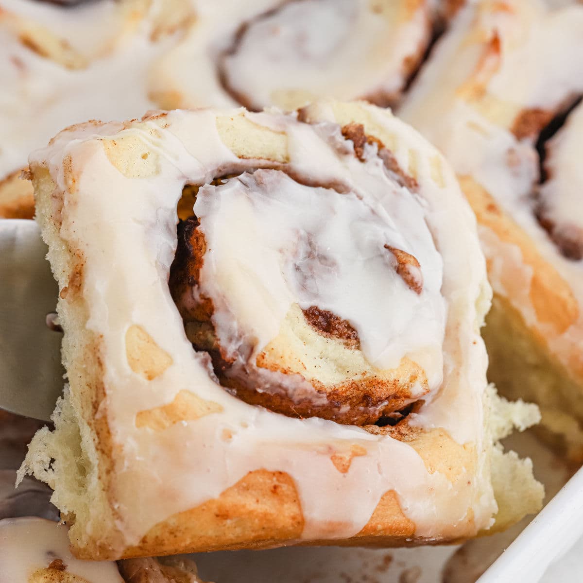 A close-up of a Paula Deen cinnamon roll being lifted from a tray, showing its swirled layers and creamy icing on top.