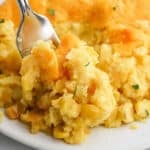 A close-up of a fork scooping Paula Deen's Corn Casserole with golden crust, visible corn kernels, and bits of fresh herbs on a white plate.