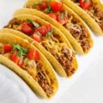 Four crunchy sloppy joe tacos filled with seasoned ground beef, shredded cheese, and topped with chopped tomatoes and fresh herbs, arranged in a white taco holder.