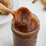 A wooden spoon scoops thick slow cooker apple butter from a glass jar. In the background, there are cinnamon sticks and part of a plate with toast spread with apple butter.