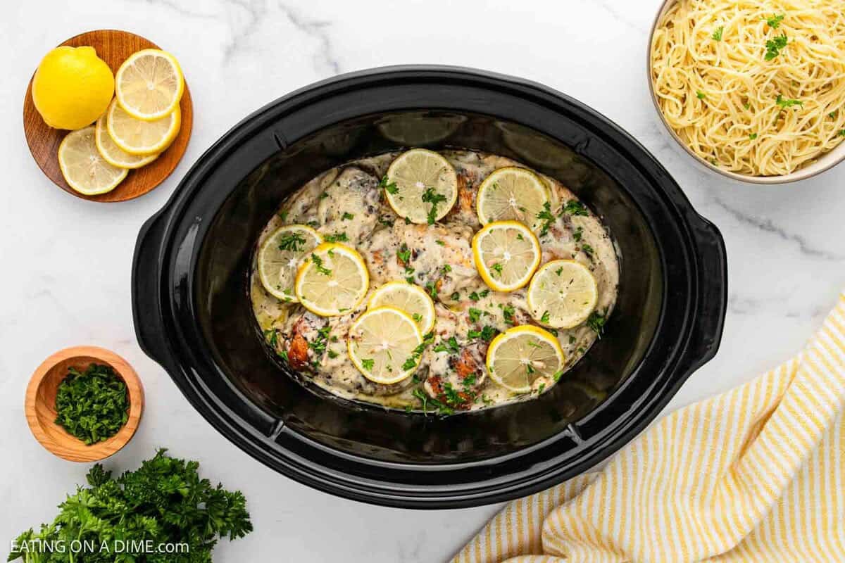 A black crock pot filled with creamy lemon chicken topped with lemon slices and parsley, surrounded by a bowl of cooked spaghetti, a plate of lemon slices, and small bowls of chopped parsley on a marble countertop.