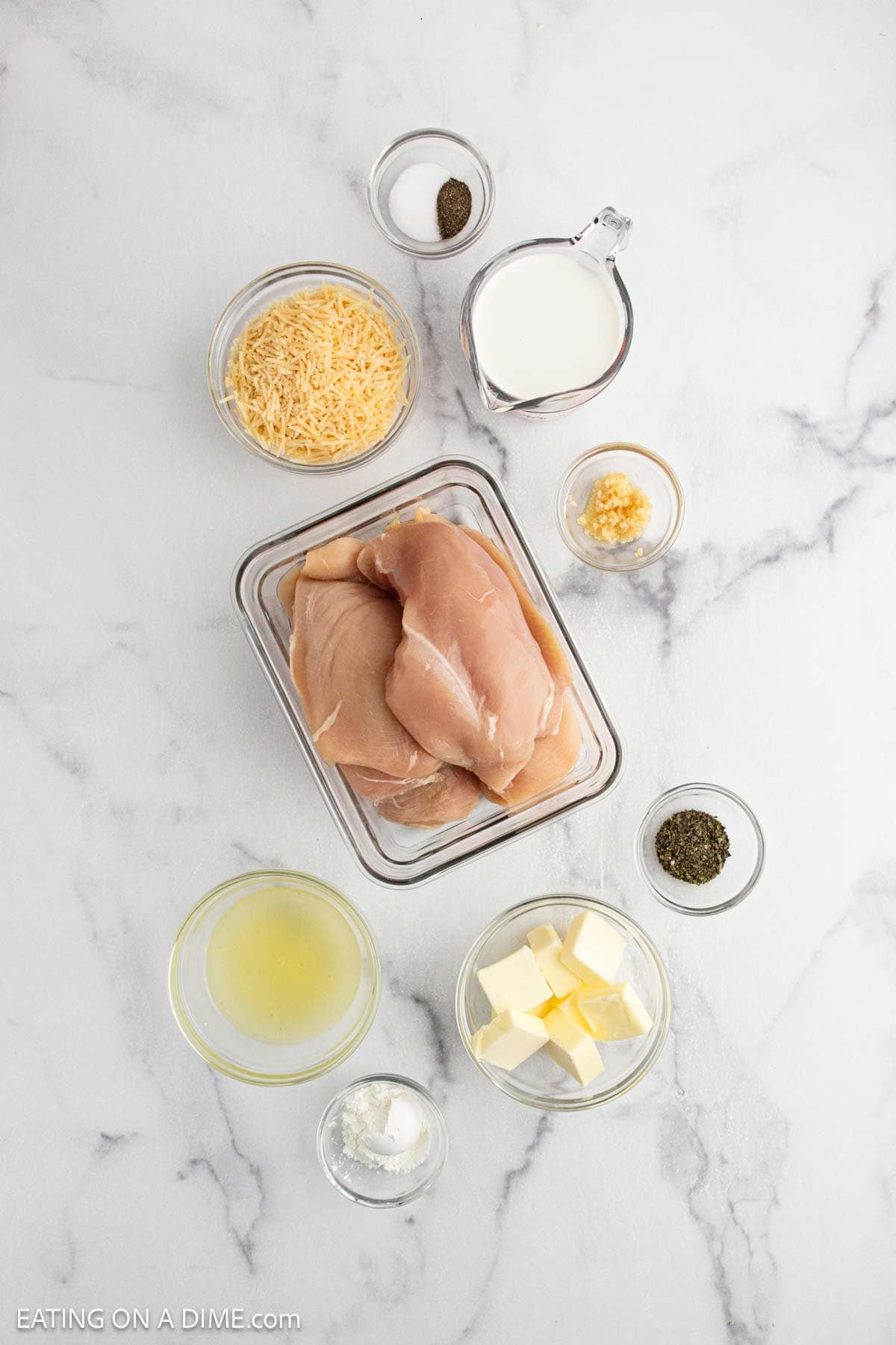 Overhead view of raw chicken breasts in a glass container surrounded by bowls of shredded cheese, milk, minced garlic, butter, chicken broth, cornstarch, dried herbs, and seasoning—perfect ingredients for crock pot lemon chicken on a white marble surface.