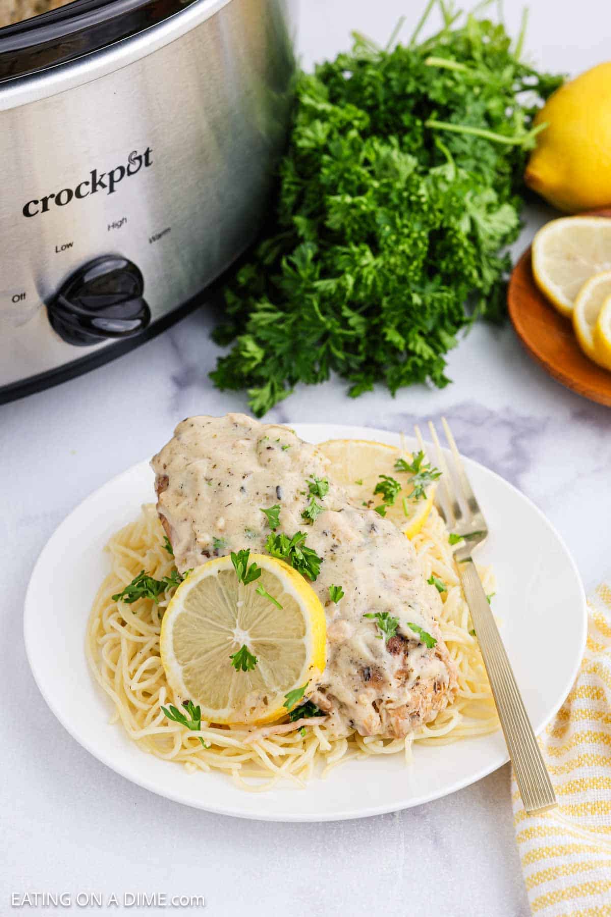 A plate of spaghetti topped with creamy crock pot lemon chicken, garnished with fresh parsley and a lemon slice. A crockpot, parsley, and sliced lemons are in the background on a marble surface.