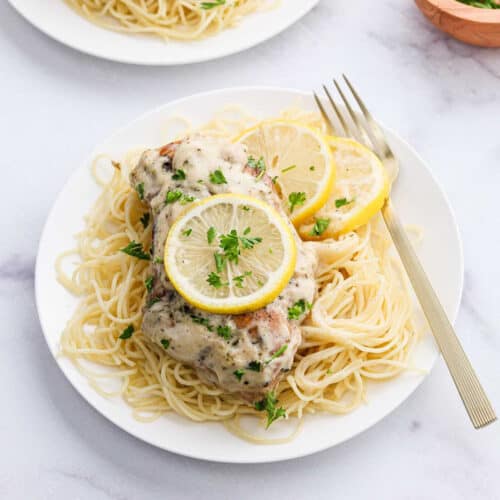A plate of spaghetti topped with creamy crock pot lemon chicken, garnished with lemon slices and chopped parsley, with a fork resting on the side of the plate.
