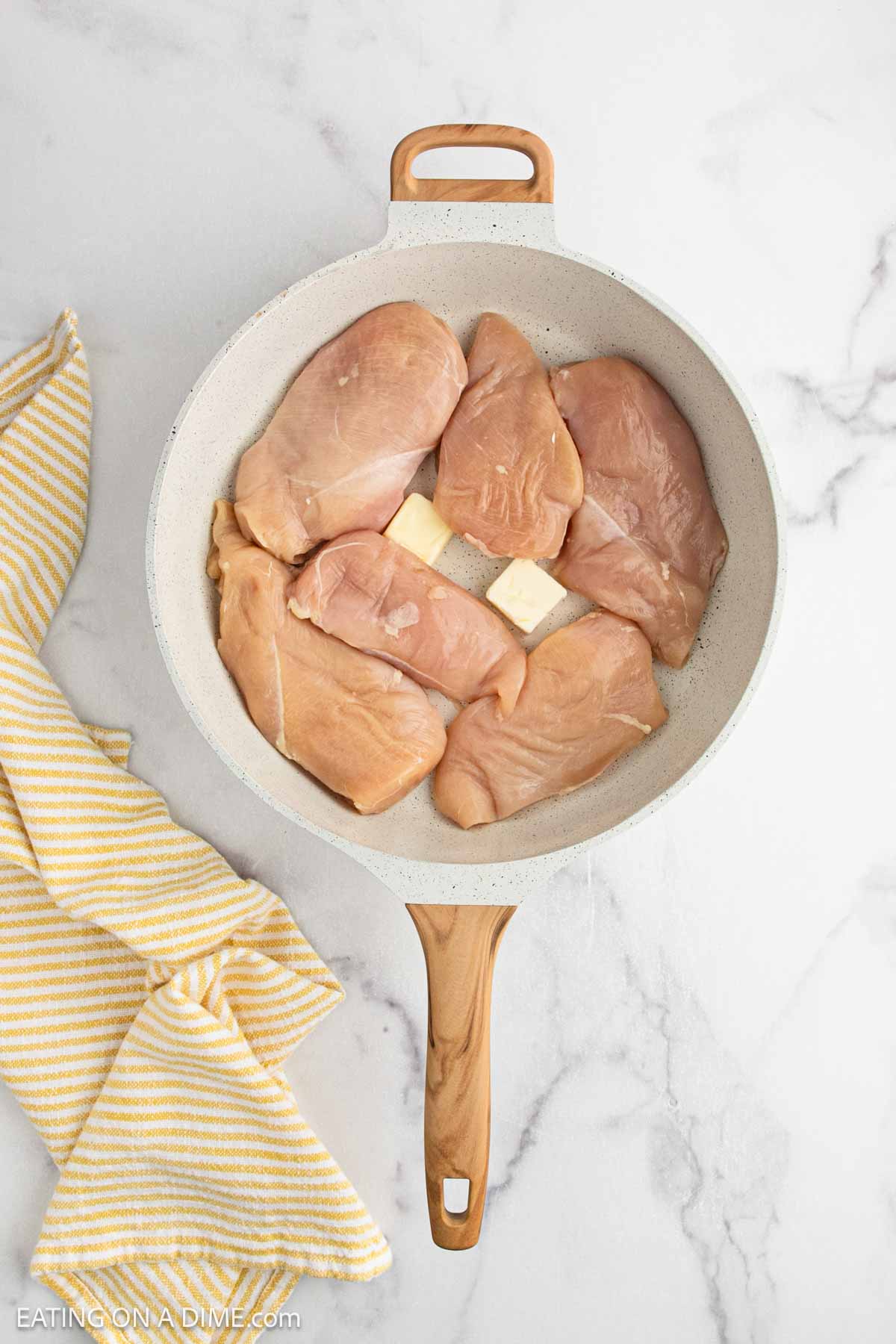 Raw chicken breasts and two butter slices in a white skillet with a wood handle, placed on a white marble surface next to a yellow and white striped kitchen towel—perfect for prepping crock pot lemon chicken.