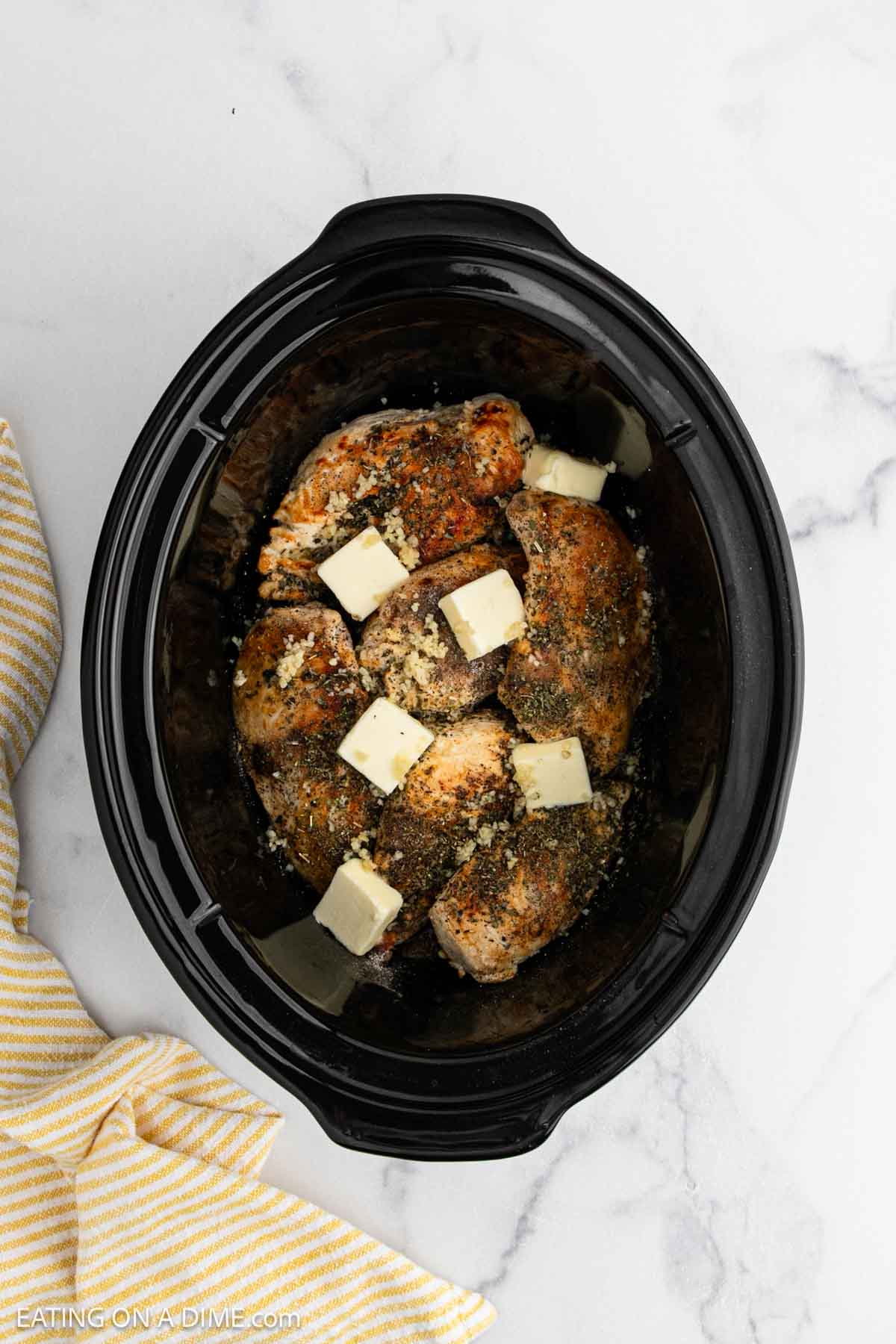 Seasoned chicken breasts in a black slow cooker, topped with butter cubes and sprinkled with herbs and spices—perfect for an easy crock pot lemon chicken recipe. A yellow striped cloth is partially visible beside the cooker on a marble surface.