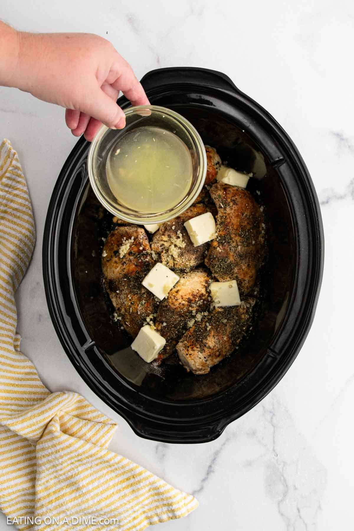 A hand pours broth from a small glass bowl over seasoned crock pot lemon chicken breasts topped with butter cubes inside a black slow cooker, with a yellow-striped cloth nearby on a marble surface.