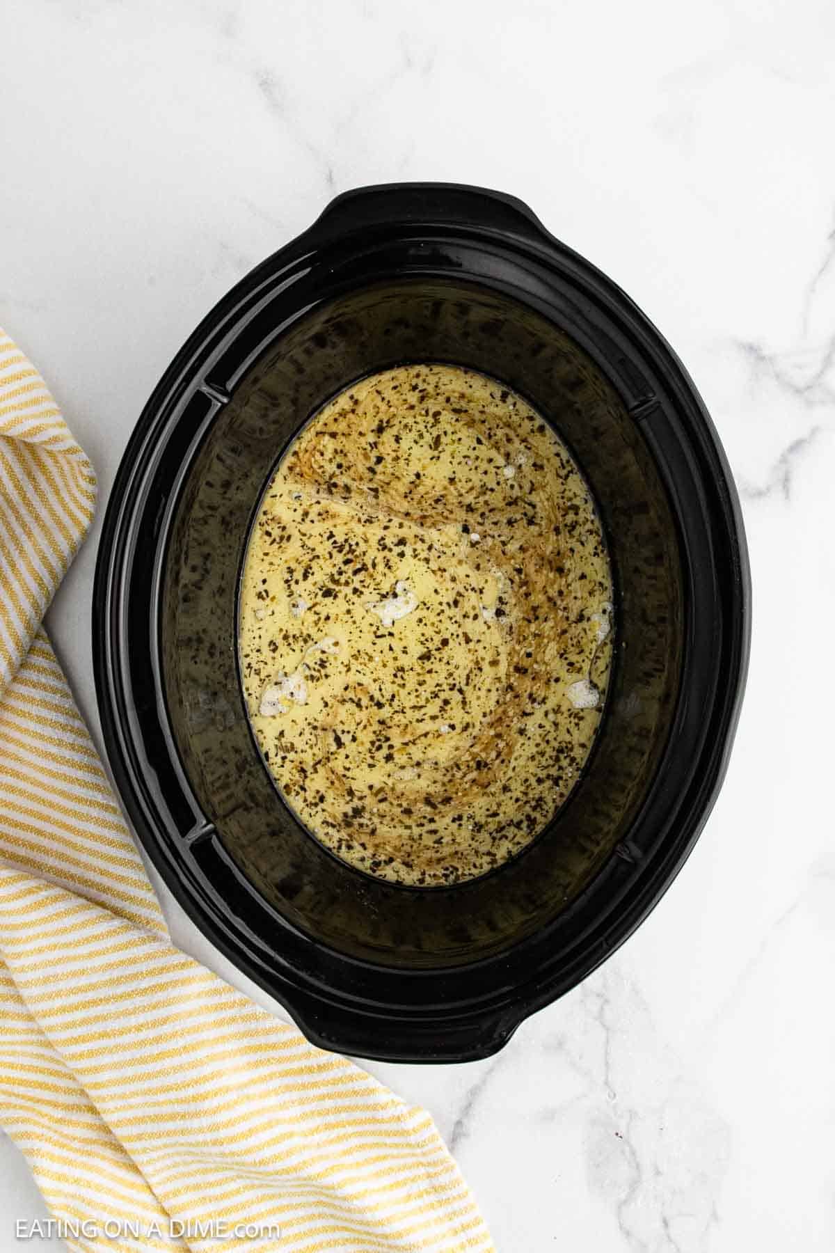 Overhead view of a black slow cooker filled with creamy crock pot lemon chicken, topped with herbs and spices. A yellow and white striped towel is placed beside the slow cooker on a white marble surface.