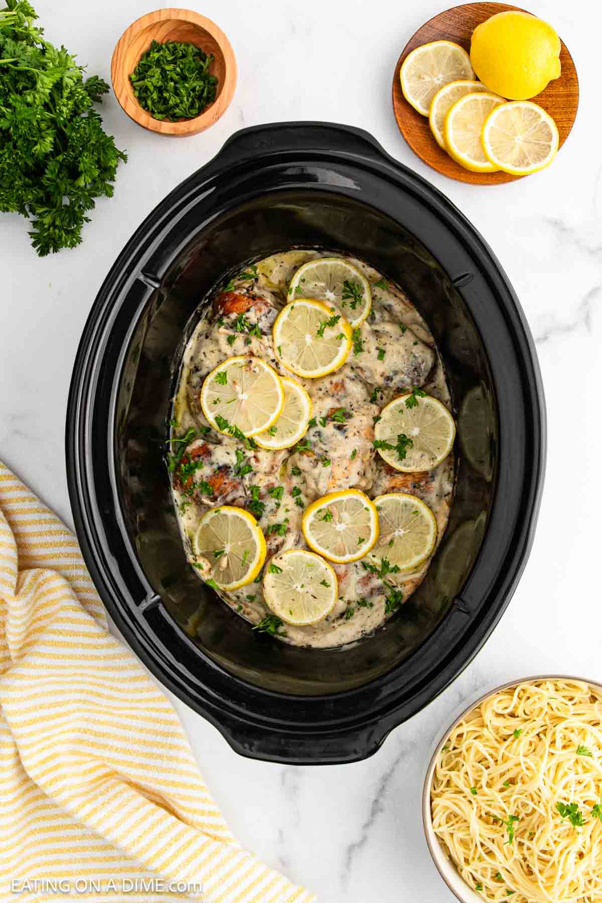 A crock pot lemon chicken dish, featuring creamy chicken breasts topped with sliced lemons and chopped parsley, sits on a white surface next to lemon slices, fresh herbs, and a plate of cooked pasta.