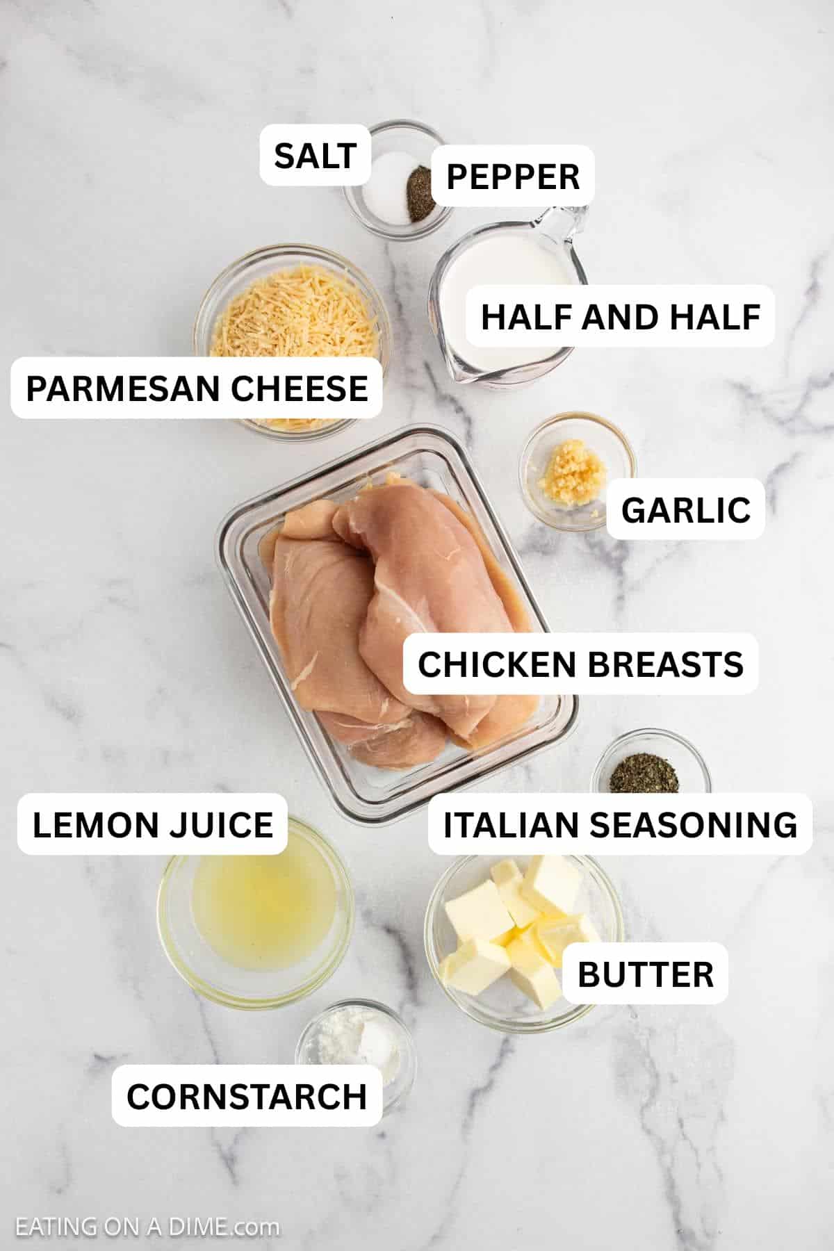 Overhead view of raw chicken breasts in a glass container surrounded by bowls of shredded cheese, milk, minced garlic, butter, chicken broth, cornstarch, dried herbs, and seasoning.